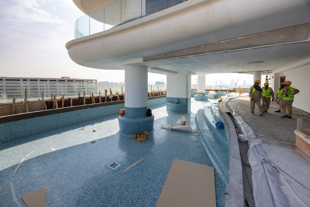 Beach Walk Residence Under construction outdoor pool area with blue tiles, workers in safety gear discussing nearby.